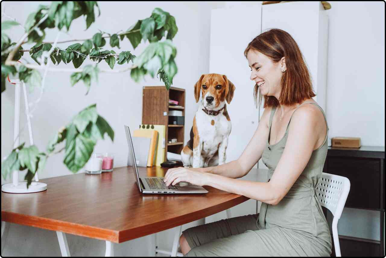 Home office setup with a woman typing on her computer and her dog sitting nearby.